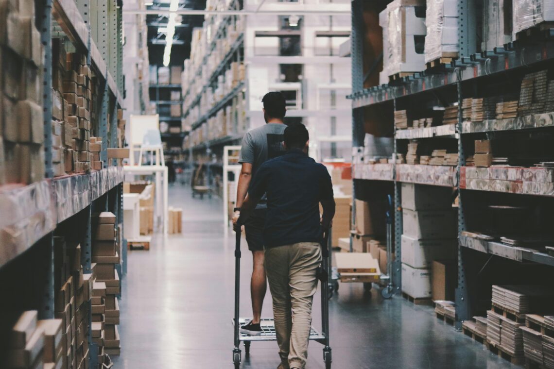 Two men navigate through a large warehouse aisle filled with stacked boxes and packaged goods. One man is riding on a flatbed cart while the other pushes it forward. The scene is well-lit with overhead industrial lighting, and the warehouse shelves are organized with various items.