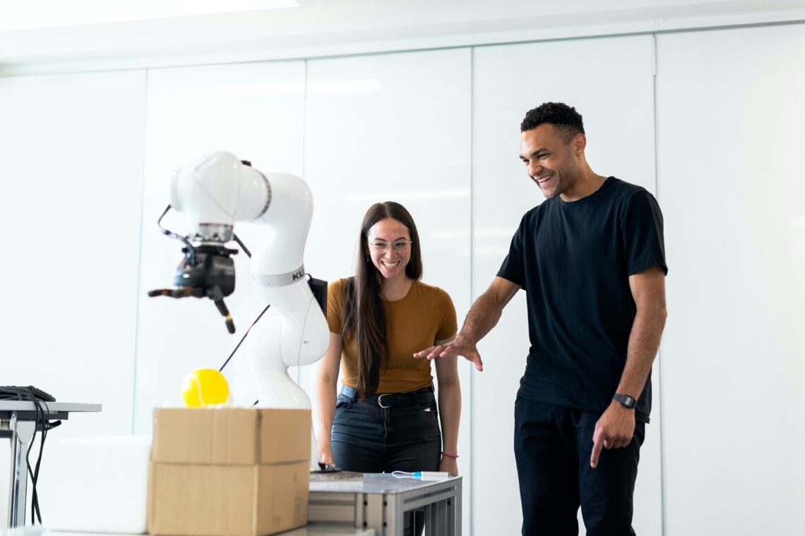 Two engineers interact with a robotic arm in a modern lab setting, smiling as they observe its functionality. The scene reflects innovation, collaboration, and the practical application of AI and robotics in technology development.
