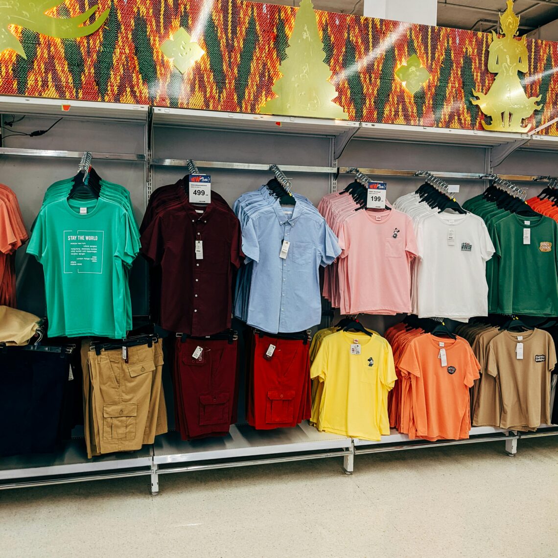 Display of neatly arranged men’s casual shirts and shorts in various colors at a retail clothing store, featuring a mix of button-ups and graphic tees under festive store decor.